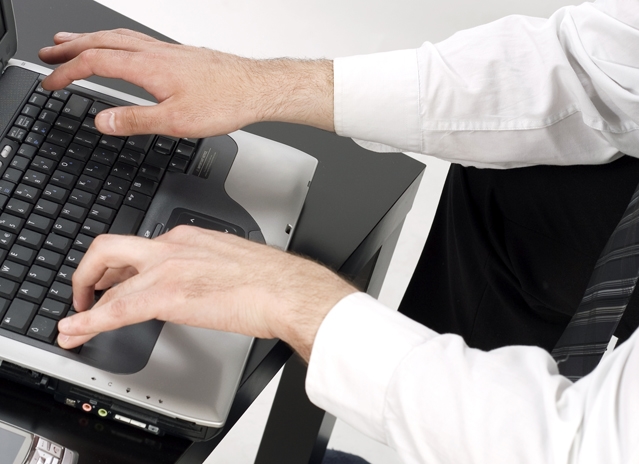 widoczna jest część klawiatury laptopa, na której coś pisze mężczyzna ubrany w białą koszulę i krawat