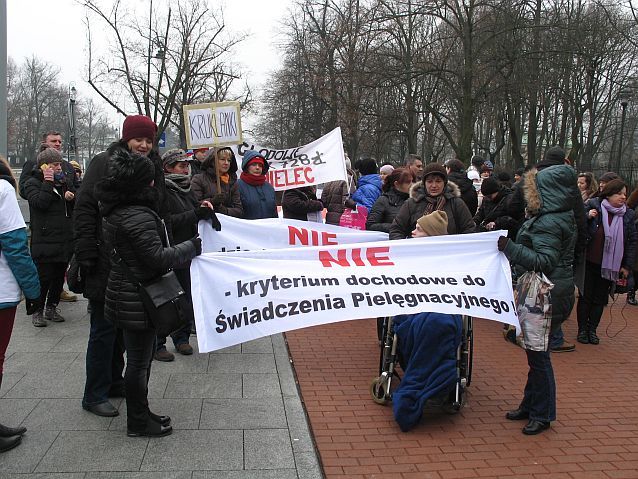 rodzice na chodniku trzymają transparent z napisem: nie kryterium dochodowe do świadczenia pielęgnacyjnego  