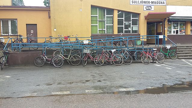 Podjazd dla wózków zastawiony przypiętymi rowerami. W tle budynek dworca PKP Sulejówek Miłosna