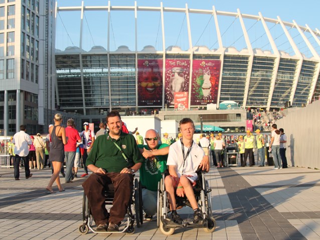 dwóch mężczyzn na wózkach i jeden mężczyzna kucający w środku pozują do zdjęcia pod stadionem w Kijowie