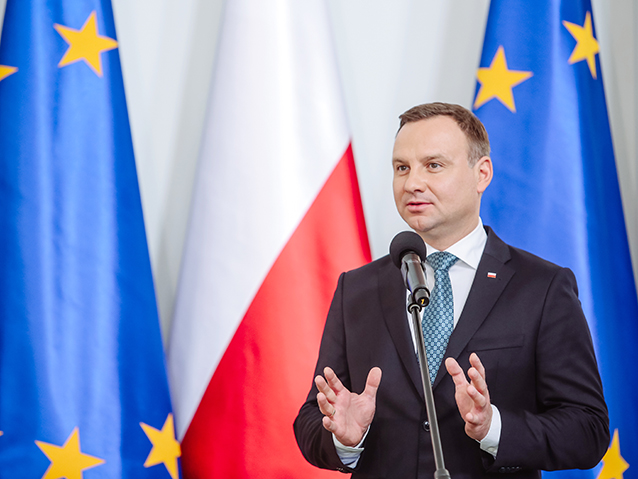 Prezydent Andrzej Duda mówi do mikrofonu na tle flag Polski i Unii Europejskiej