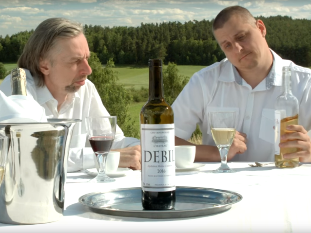 dwóch mężczyzn siedzi przy stole, na środku stoi wino z etykietą Debil