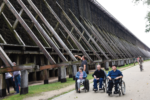 trzy osoby na wózkach przy drewnianej konstrukcji przypominającej dach 