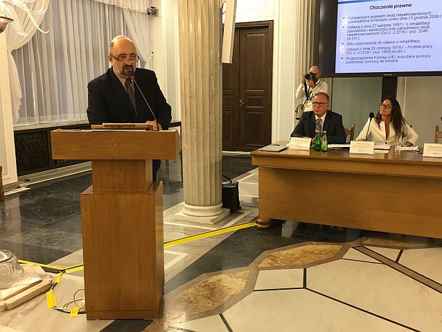 Mirosław Przewoźnik stoi przy mównicy przed mikrofonem, w tle dwie osoby za stołem prezydialnym