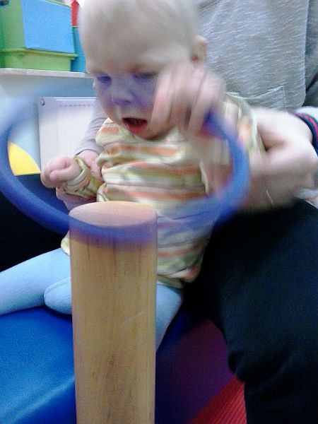 Dziecko podczas terapii próbuje nałożyć z pomoca terapeuty plastikową obręcz na drewniany kołek