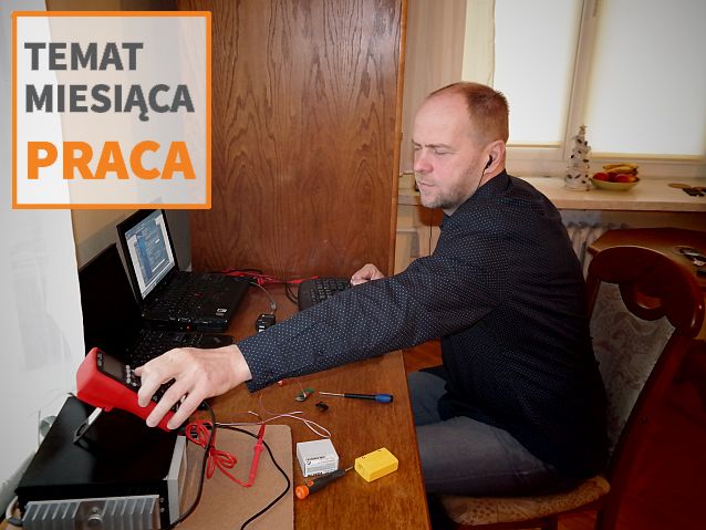 Jan Szuster siedzi przy biurku, na którym stoją dwa laptopy, i sięga do elektronicznego miernika. Na zdjęciu napis: Temat miesiąca: Praca