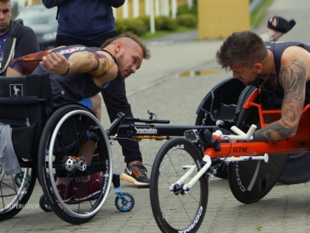 dwóch umięśnionych mężczyzn na wózkach, schylających się. z prawej: Krzysztof Stern