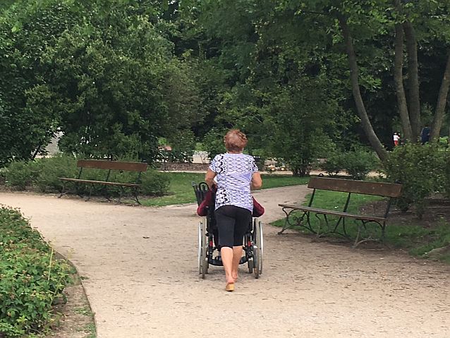 drogą po parku idzie kobieta, prowadzaca osobę na wózku