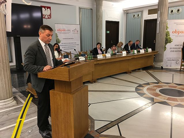 Krzysztof Kaca przemawia stojąc przy mównicy, w tle kilka osób za stołem prezydialnym