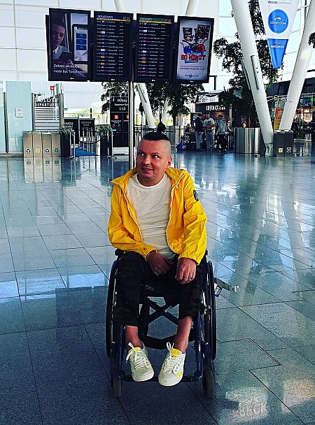 Bartek Skrzyński siedzi na wózku na środku hali portu lotniczego, w tle elektroniczne tablice przylotów i odlotów