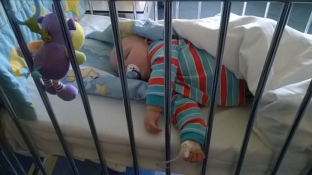 Małe dziecko śpi w łóżeczku szpitalnym. Na pierwszym planie wisi maskotka, rączka dziecka podłączona jest do rurki