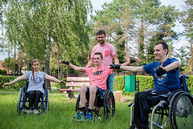 Troje młodych ludzi na wózkach ćwiczy na świeżym powietrzu, jednemu z nich pomaga rehabilitant