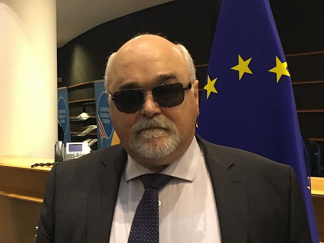 Yannis Vardakastanis w czarnych okularach stoi na tle flagi Unii Europejskiej