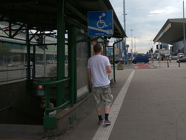 Mężczyzna stoi na ścieżce prowadzącej dla osób niewidomych, tuż nad głową ma znak informujący o rampie dla osób na wózkach