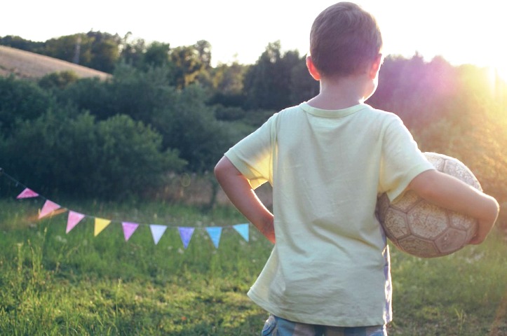 chłopiec stoi tyłem, trzyma piłkę do nogi pod pachą, w okół jest zielono, krajobraz przypomina wieś