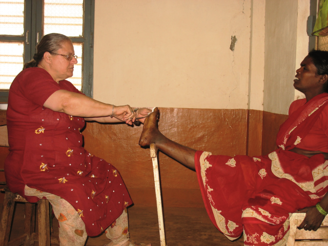 Helena Pyz w skupieniu opatruje palca stopy kobiety z indii w średnim wieku, której noga jest podparta na specjalnym białym kiju