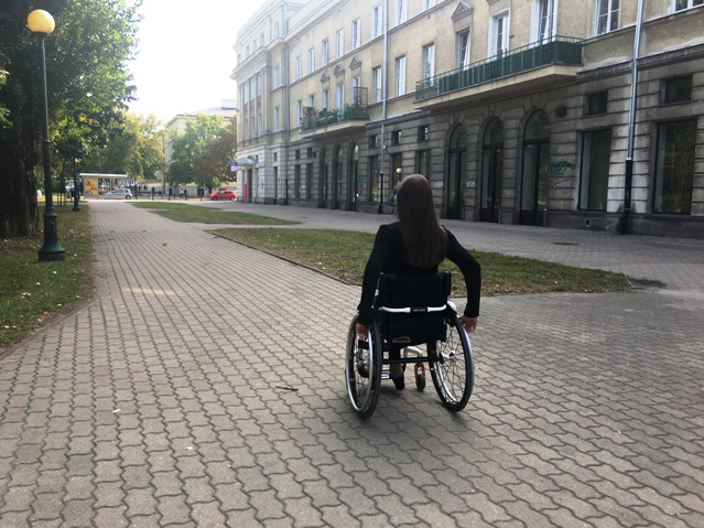 kobieta ubrana na czarno porusza się na wózku po chodniku na warszawskim Muranowie