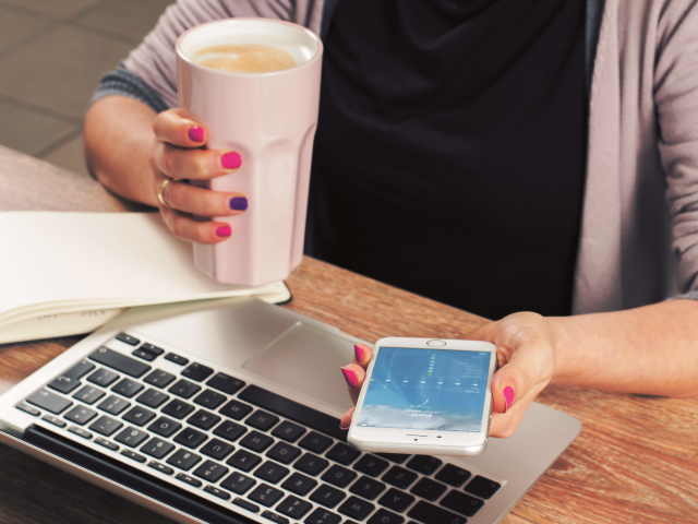 Kobieta siedzi przed laptopem, w jednej dłoni trzyma kawę, w drugiej telefon
