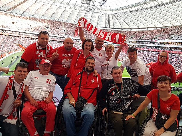 Kibice z niepełnosprawnościami, w tym na wózkach, oraz ich asystenci pozują w polskich barwach do zdjęcia na tle wypełnionego stadionu