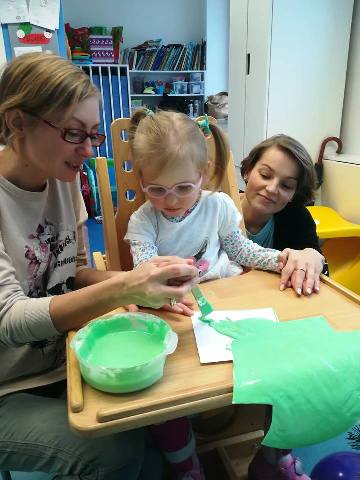 Jedna nauczycielka pokazuje dziewczynce jak pokrywać zieloną farbą wyciętą z papieru choinkę. Za nimi przez raimię dziecka zagląda prawdopodobnie mama, ponieważ trzyma dziecko za dłoń