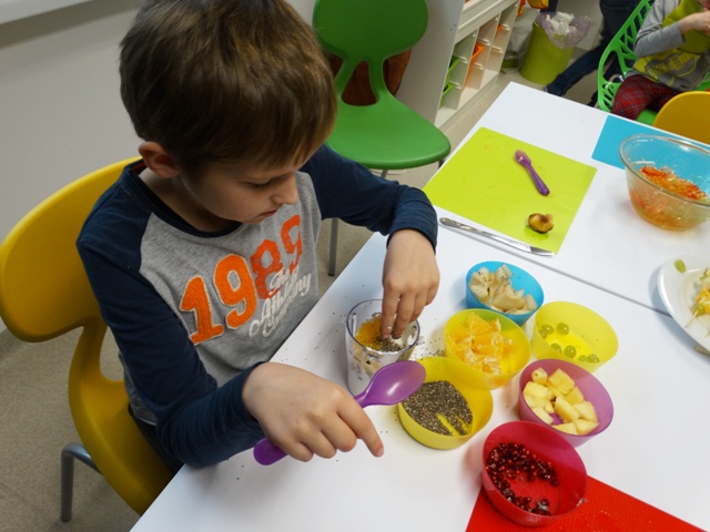 Chłopiec wsypuje do szklanki płatki, mandarynki i ziarnka. W drugiej dłoni trzyma fioletową łyżkę