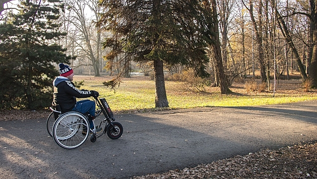 Mężczyzna jedzie przez park ręcznym wózkiem inwalidzkim kierując podłączoną z przodu elektryczną przystawką napędzającą całość