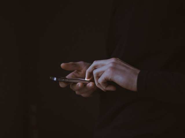 osoba ubrana w czarny sweter lub bluzę w jednej dłoni trzyma telefon, drugą wystukuje coś na nim