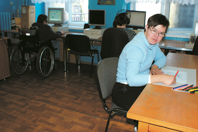 młoda kobieta z krótkimi włosami i z okularami na twarzy maluje przy biurku, za nią w tle siedzą dwie osoby, korzystające z komputerów