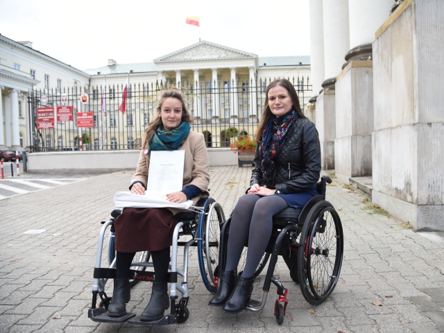 Hanna Gospodarczyk, siedząc na wózku trzyma w dłoniach dyplom. Obok niej młoda kobieta, także na wózku. Kobiety znajdują się przed bramą ratusza