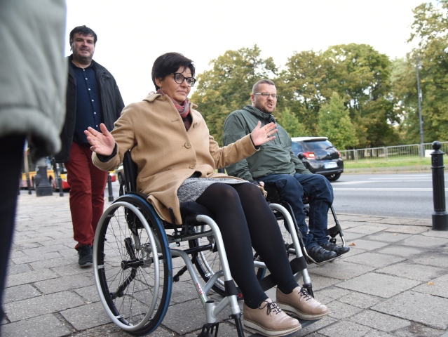 Justyna Glusman siedząc na wózku, rozkłada ręce, obok niej mężczyzna z niepełnosprawnością poruszający się na wózku, a za kandydatką na prezydenta Warszawy asystent Integracji