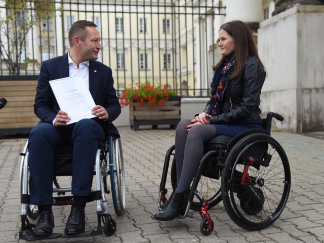 Paweł Rabiej, siedząc na wózku, trzyma w dłoniach Dyplom zdania Testu Integracji wiedzy o niepełnosprawności i przejazdu z Integracji do Ratusza. Mężczyzna patrzy na młodą kobietę na wózku. Uśmiechają się.
