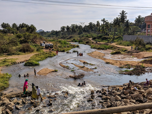 Płytka rzeka, stojące na kamieniach 3 kobiety, w oddali samochody na brzegu, palmy i zarośla po obu stronach