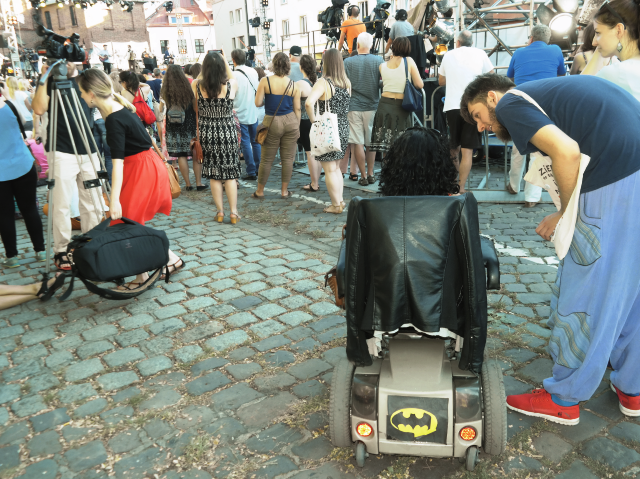 plac, na którym znajduje się tłum ludzi. wśród tłumu jest na wózku kobieta na wózku elektrycznym, która rozmawia z nachylającym się do niej chłopakiem