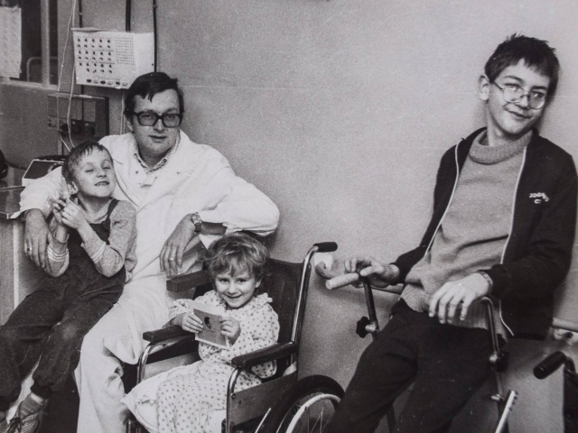 doktor Piotr Janaszek w białym kitlu obok niego trójka uśmiechniętych dzieci, jedno na kolanach, dwoje na wózku
