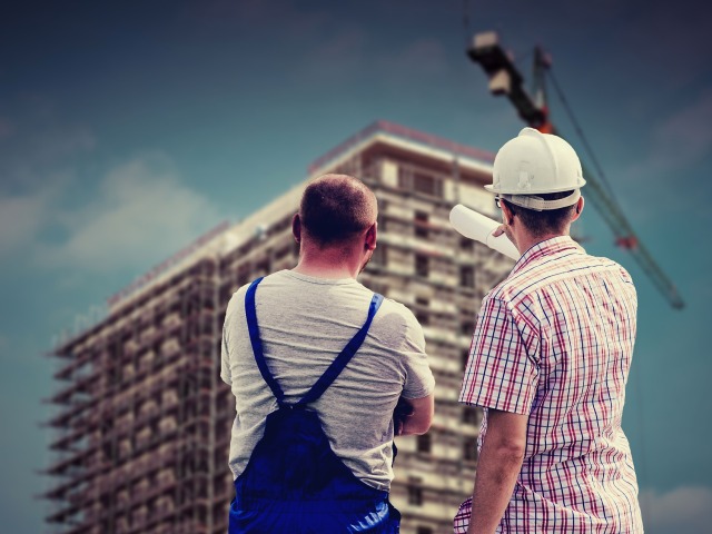 dwóch mężczyzn jeden w roboczym ubraniu drugi w kasku stoją tyłem przed nimi blok w budowie