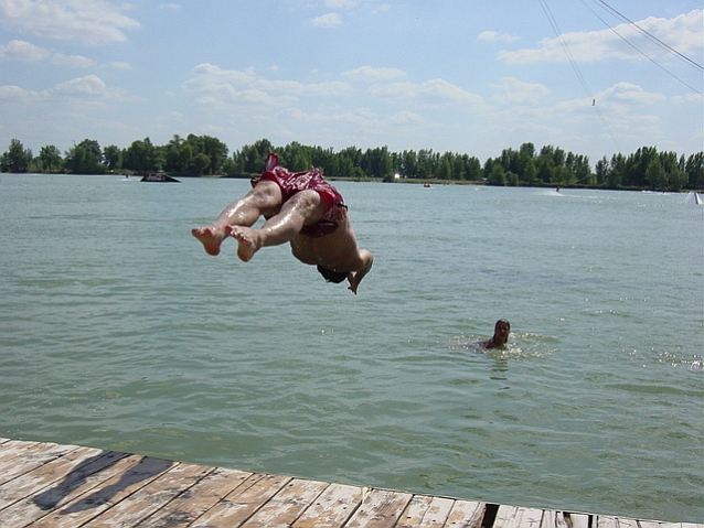 Mężczyzna skacze z pomostu do wody, odwrócony do niej plecami, w wodzie jest inna osoba