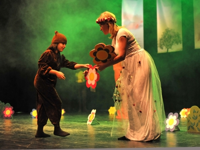 teatr z Macedonii na scenie kobieta ubrana w zwiewną suknię i wianek wręcza dziewczynce w czarnym kostiumie i czapce na głowę kwiat z papieru