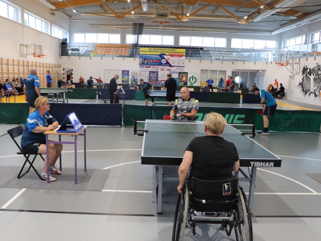 kobieta i mężczyzna na wózkach przy stole tenisowym grają z lewej strony siedzi kobieta przy stoliku sędzia