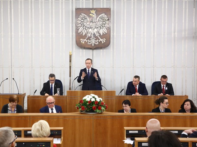 Tomasz Grodzki przemawia na mównicy w senacie nad nim wisi godło Polski senatorowie siedzą po bokach