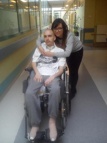 Jakub chmielowiec siedzi na wózku na szpitalnym korytarzu za nim przytulona do niego jego narzeczona