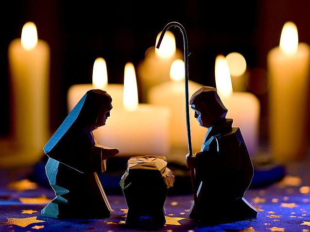 Drewniane figurki Marii i Józefa klęczących nad Jezusem w żłóbku. W tle płonące świece
