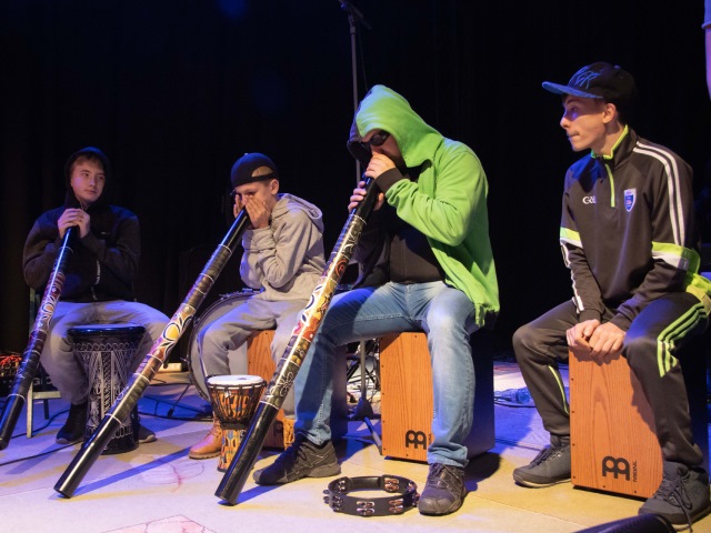 trzech chłopców i jeden mężczyzna na scenie grają na podłużnych kolorowych tubach przed nimi bębny i tamburyn