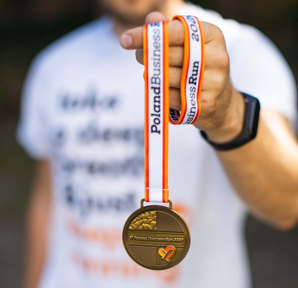 mężczyzna trzyma w ręku medal biegu złoty krążek na biało pomarańczowej wstędze