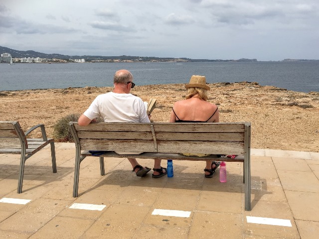 starsi kobieta i mężczyzna siedzą tyłem na ławce przed nimi morze, mężczyzna czyta książkę