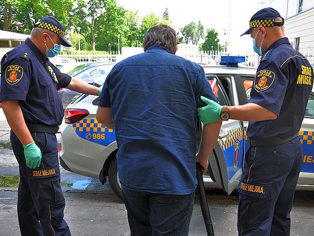 Dwóch strażników miejskich pomaga starszemu człowiekowi o kuli wsiąść do radiowozu straży miejskiej