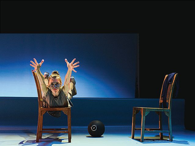 W teatrze na scenie stoją dwa krzesła, na jednym z nich leży na brzuchu kobieta, wyciągając ręce w stronę widza