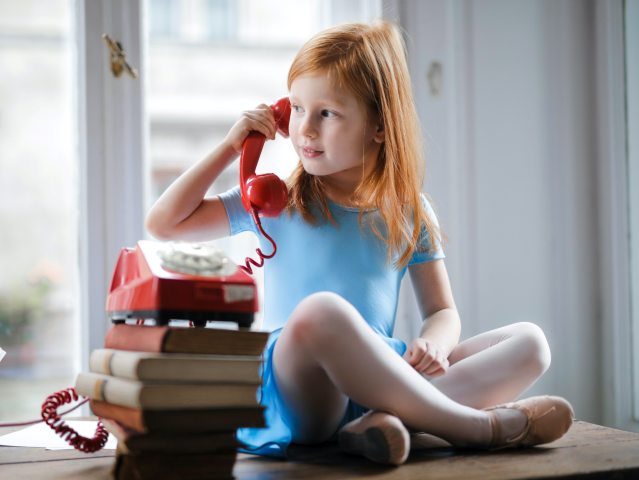 kilkuletnia dziewczynka siedzi po turecku na stole i rozmawia przez słuchawkę czerwonego telefonu