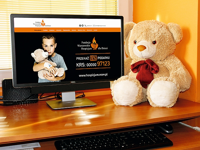 Monitor komputera z otwartą stroną Warszawskiego Hospicjum dla Dzieci. Obok monitora pluszowy miś