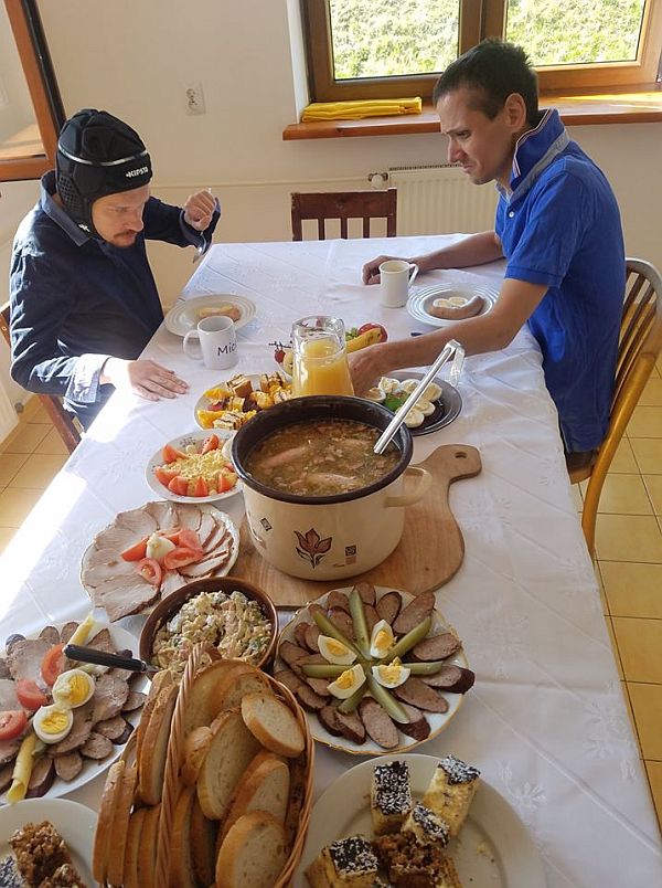 Dwóch mężczyzn siedzi przy stole, zastawionym wielkanocnymi potrawami, na pierwszym planie garnek żuru z kiełbasą