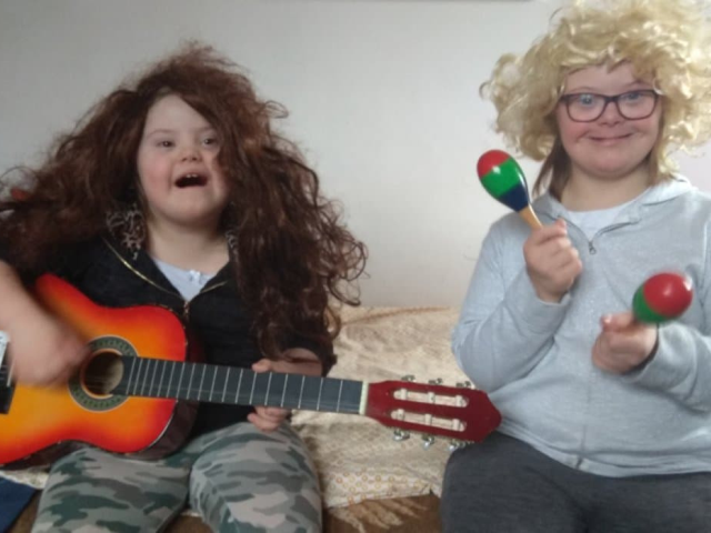 dwie dziewczynki z zespołem downa w perukach jedna gra na gitarze a druga trzyma w ręku grzechotki